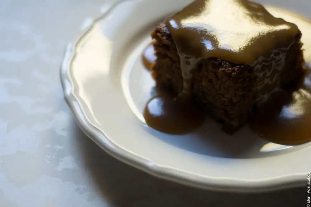 Sticky Toffee Pudding Κέικ με Χουρμάδες και Σάλτσα Καραμέλας Βουτύρου www.thefoodiecorner.gr Photo description: A piece of sticky toffee pudding with sauce all over it and down the sides, sitting on a white plate, light coming from behind darkening the front of the cake and making the sauce shine on top.