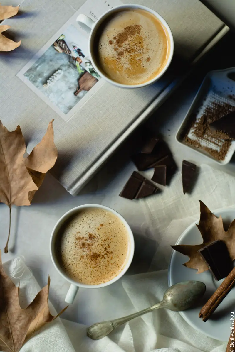 Food Styling and Photography Retreat – Crete, Greece 2017 www.thefoodiecorner.gr Photo description: Two cups of coffee on a marble like background, one sitting on a similar coloured book. Around them dry autumn leaves, some pieces of dark chocolate and a grater, and a cinnamon stick on a saucer.