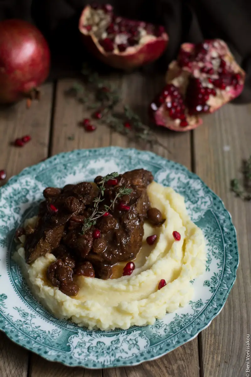 Slow Cooker Pomegranate and Tomato Beef with Sultanas www.thefoodiecorner.gr Photo description: Three quarter view of a plate of beef over mashed potatoes with some pomegranate arils sprinkled on top. The antique plate has a pretty design around the edge and is sitting on a wooden background. Behind the plate are some pomegranates, one of them broken open to reveal the arils inside. A couple of sprigs of thyme decorate the meat.