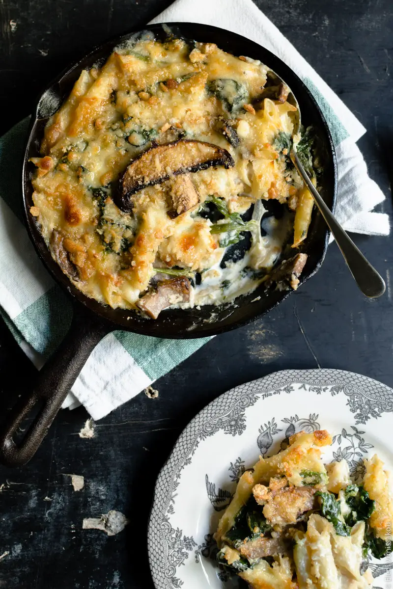 Pasta Bake with Spinach and Portobello Mushrooms in Bechamel Sauce www.thefoodiecorner.gr Photo description: To the top of the photo, the skillet with the pasta bake, a spoon lying inside and a piece missing. To the bottom right, a plate with a pretty pattern round the edge, with a messy piece of pasta bake on it. Some penne coated with creamy béchamel are visible, as well as some spinach and pieces of golden crunchy topping.