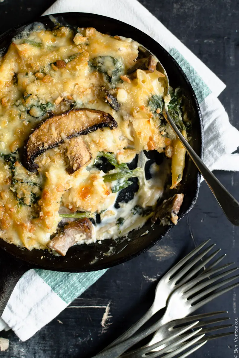 Pasta Bake with Spinach and Portobello Mushrooms in Bechamel Sauce  www.thefoodiecorner.gr Photo description: A close up of the skillet with a spoon lying in it. A piece of pasta bake has been taken out and some pieces of spinach, mushrooms and creamy bechamel are visible from within. To the bottom right a bunch of forks. The skillet is on the folded tea towel.