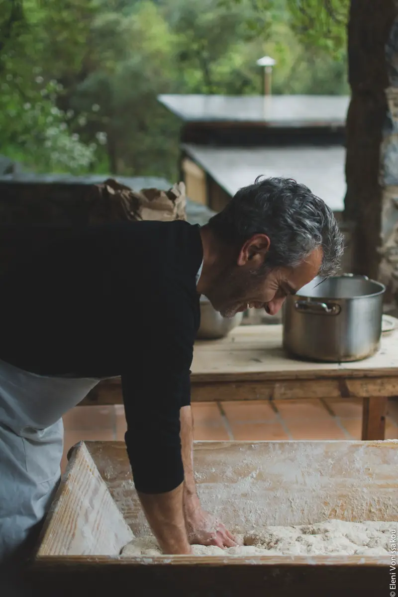 Miliaworkshop2017 www.thefoodiecorner.gr photo description: Side view of a man, apron on, sleeves rolled up, bending over a large wooden basin filled with dough. In the background some trees and the roof of an outbuilding. 