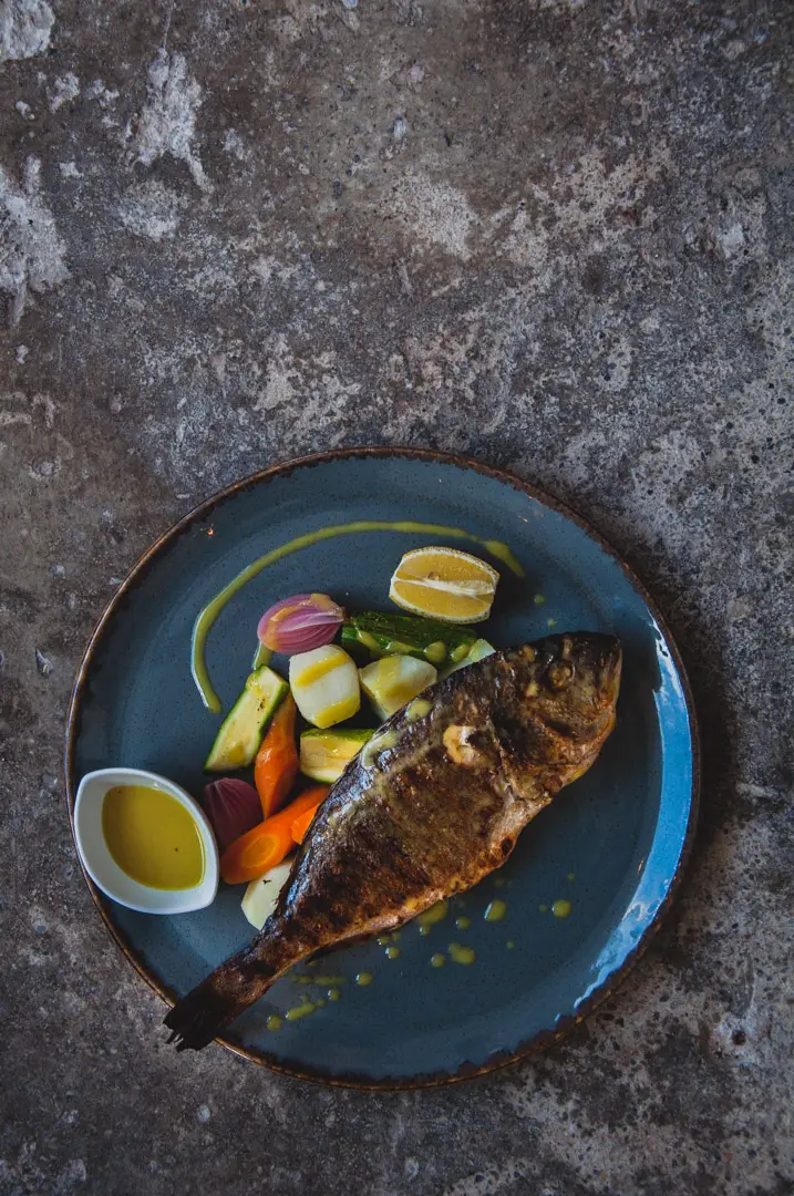 Miliaworkshop2017 www.thefoodiecorner.gr Photo description: a whole, cooked fish lying across a round plate. Underneath it, some boiled potatoes, courgettes and carrots, a wedge of lemon and a small bowl of yellow sauce. Plate is on a rough concrete surface.