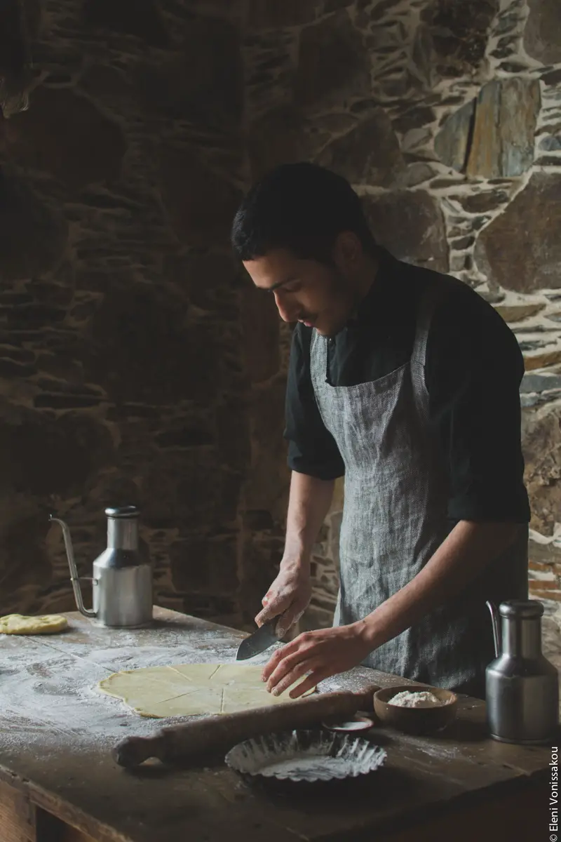 Miliaworkshop2017 www.thefoodiecorner.gr Photo description: A ¾ side view of a young man cutting some rolled out pastry. The wooden table is covered in flour and on it is a rolling pin and some metal canisters. The man is standing in front of a stone wall.