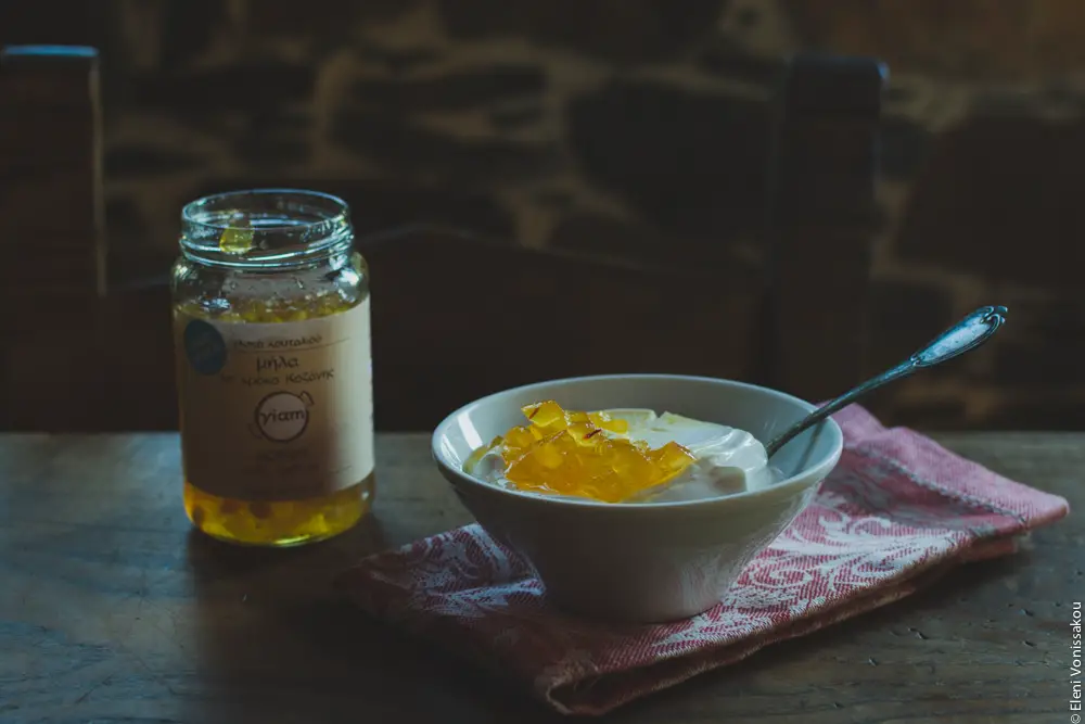 Milia Workshop 2017 – A long overdue recap. Part 1. www.thefoodiecorner.gr - Photo description: A bowl of yoghurt with a dollop of yellow, jam like “spoon sweet” on top. A spoon is sitting in the yoghurt. To the left, the jar of spoon sweet. The bowl is sitting on a folded napkin with a pretty pattern, all on a wooden table. In the background a stone wall is visilble.
