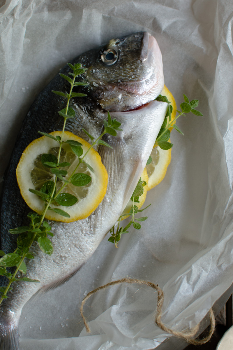 Gilt Head Bream (Dorade) – How to cook a whole fish in the slow cooker www.thefoodiecorner.gr Photo description: a close view of a raw gilt head bream, lying on a piece of baking paper prepped for cooking. There are some lemon slices barely visible, protruding from the cavity together with some fresh oregano sprigs. Some more lemon and oregano sprigs decorate the top of the fish. 