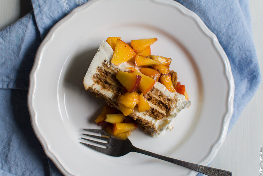 Greek Yoghurt, Mascarpone and Speculoos (Biscoff) Cookie Fridge Cake with Peaches www.thefoodiecorner.gr Photo description: A slice of the cake on a pretty plate, with peaches spooned over the top. The layers of biscuit and cream are visible under the peach. A fork also lies on the plate, which is sitting on a linen tea towel.