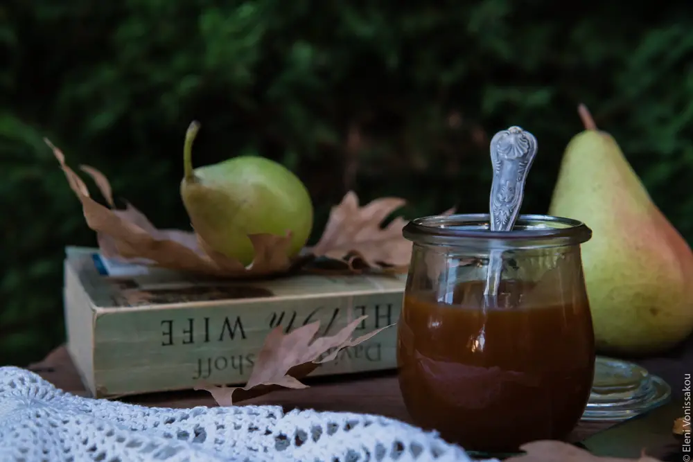 Easy, No Mixer Apple Cake with Salted Caramel Sauce www.thefoodiecorner.gr Photo description: Side view of a small jar of caramel sauce on a wooden table with a pretty spoon in it. Just behind it and to its left is a worn book with some brown leaves on it and a pear. Another pear is in the background.