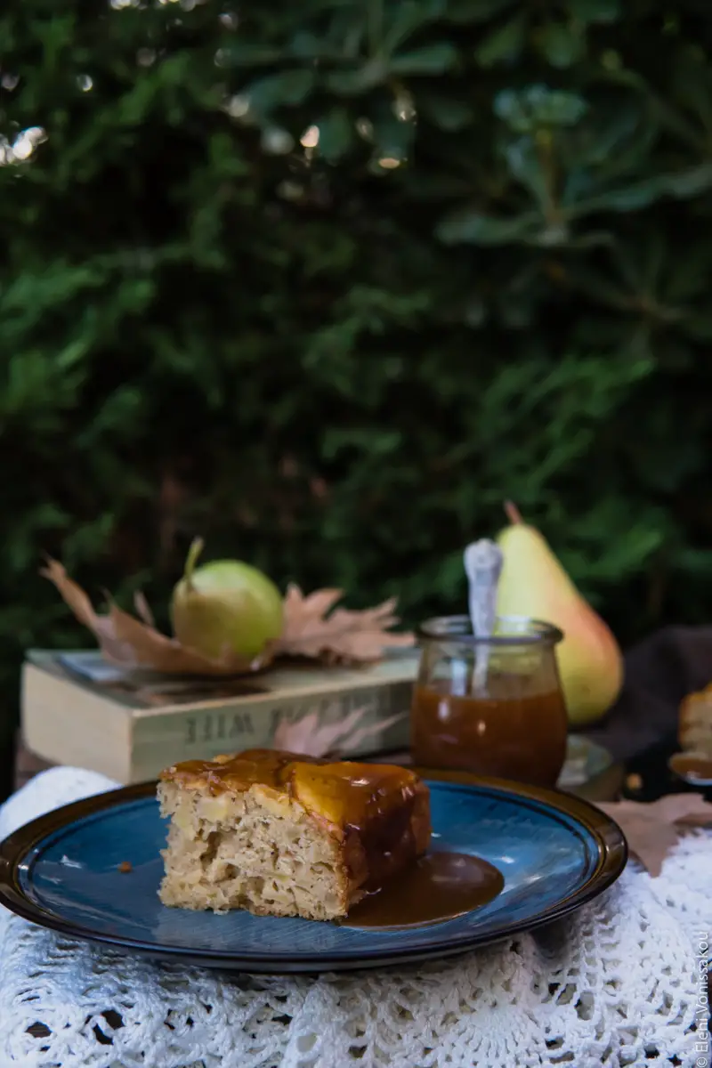 Easy, No Mixer Apple Cake with Salted Caramel Sauce www.thefoodiecorner.gr Photo description: A side view of a piece of cake on a plate. The inside of the cake is facing us and there is some caramel dripping over the other side. In the background a book, some pears and some brown leaves. All against a backdrop of dense foliage.