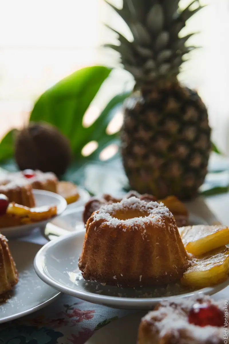 Greek Ravani Turned Mini Pina Colada Cakes www.thefoodiecorner.gr Photo description: A side view of a cake, its plate sitting on top of other plates. A whole pineapple is visible against a bright background, as is part of a monstera leaf.