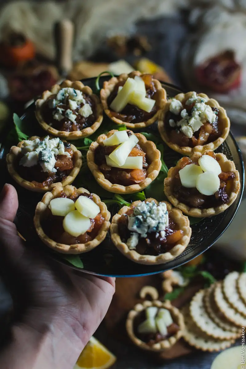 Savoury Tarts with Pear Chutney and Blue Cheese or Gruyere www.thefoodiecorner.gr Photo description: A hand entering the frame from the left side, holding a plate of tarts. Barely visible in the background is the rest of the cheese board.
