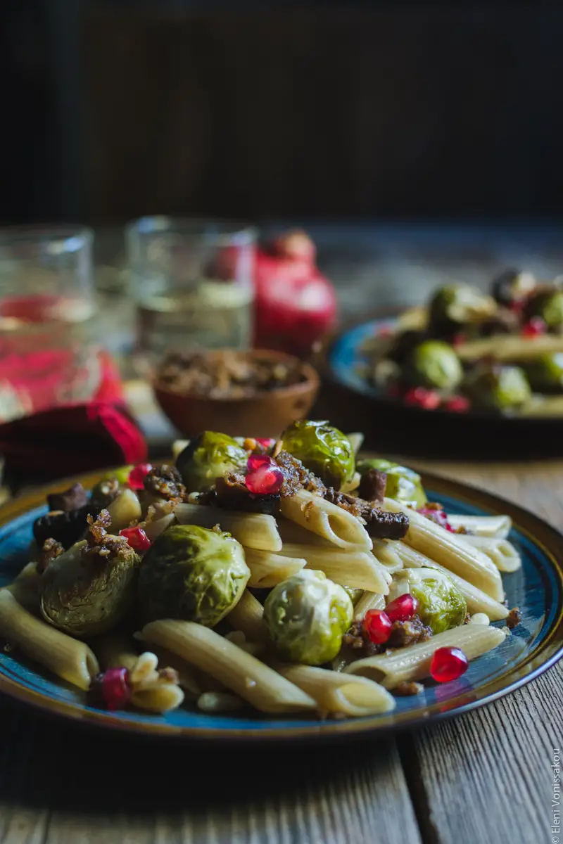 Pasta with Balsamic Roasted Brussels Sprouts and Shiitake Mushrooms (Vegan) www.thefoodiecorner.gr Photo description: A side view of a plate of pasta, dotted with Brussels sprouts and mushrooms, and sprinkled with pomegranate arils. In the dark background barely visible are the second plate, two glasses of wine and the whole pomegranate.
