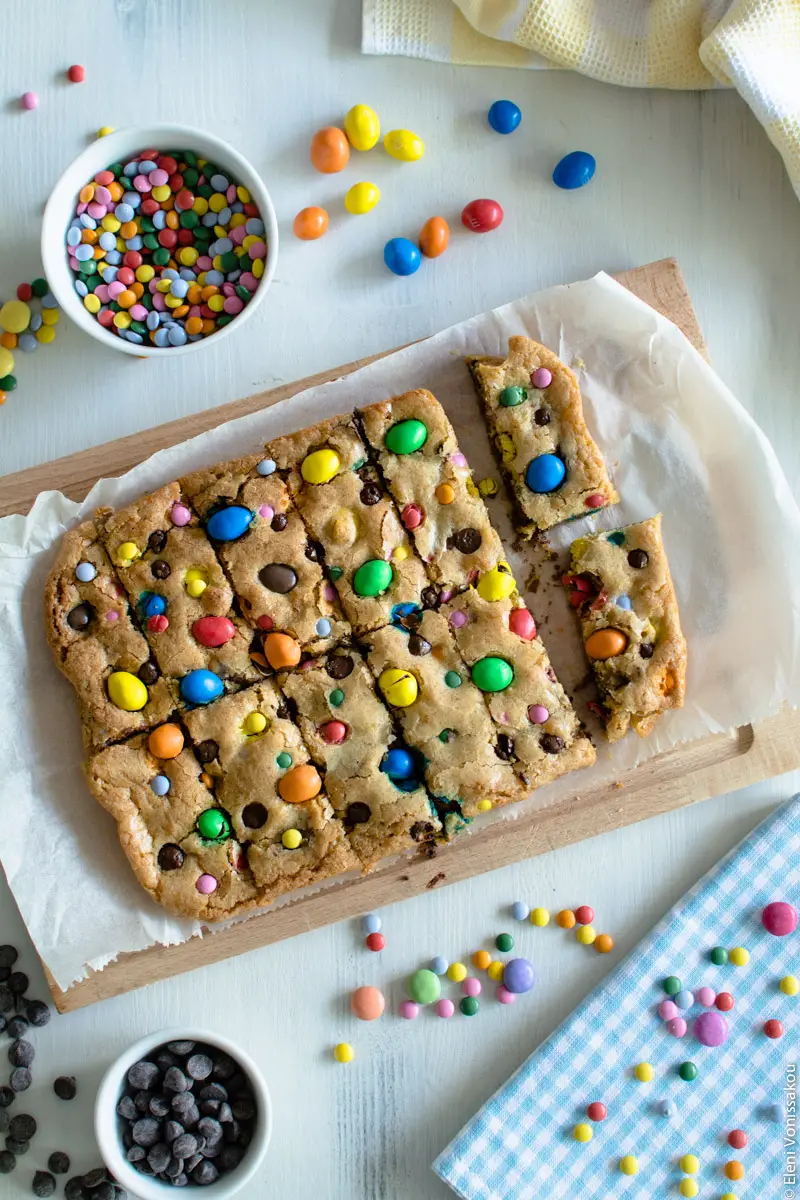 Easy, One-bowl, Colourful Chocolate Chip Cookie Bars www.thefoodiecorner.gr Photo description: The cookie bars on the chopping board, freshly cut but not yet separated. Around the board are some scattered candies on the wooden surface and in a bowl, and some chocolate chips also on the surface and in a small bowl. In the top right and bottom right corners are a couple of colourful, chequred tea towels.