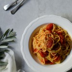 Σπαγγέτι με Ντοματίνια και Ελαιόλαδο www.thefoodiecorner.gr. Spaghetti cooked with cherry tomatoes, served on a pretty white plate. Plate with fork and spoon sitting on a marble surface with a few branches from an olive tree and a muslin cloth to decorate.