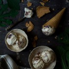 Two little handmade ceramic bowls with ice cream, a linen tea towl, some fig tree leaves and a couple of ice cream cones, one of them broken into pieces, arranged on a dark surface. Anthotyro Cheese, Honey, and Balsamic Baked Fig Ice Cream www.thefoodiecorner.gr