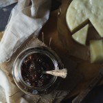 Slow Cooker Peach and Beetroot Chutney www.thefoodiecorner.gr Photo description: A dark and shadowy photo with an overhead view of some dark brown coloured chutney in a pretty jar, a silver spoon sticking out, sitting on a wooden bread board with a round of yellow cheese next to it. A bit of muslin is draped vertically along the board and the jar is sitting on some squares of burlap fabric.