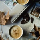 Homemade Pumpkin Spice Latte www.thefoodiecorner.gr 01 Photo description: Two cups of pumpkin spice latte with a sprinkling of spices on top, one of them sitting on a white marble-like surface, the other on a grey coloured cookery book. Cups are surrounded by dry autumn leaves, pieces of dark chocolate and a chocolate grater. At the bottom right of the photo is a saucer with sticks of cinnamon, a dry leaf and a piece of chocolate on it, and an old silver spoon propped up against it, sitting on a piece of off-white muslin cloth.