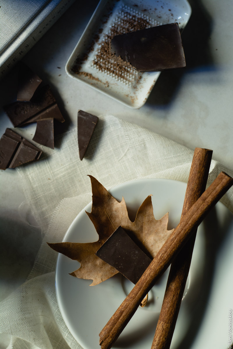 Pumpkin Spice Latte, Καφές Λάτε με Γεύση Κολοκύθας και Μπαχαρικών www.thefoodiecorner.gr 04 Photo description: A closer look at the small white porcelain chocolate grater surrounded by pieces of dark chocolate, and the saucer with two large sticks of cinnamon lying across it. A dry leaf and a piece of chocolate are also on the saucer. Photo has dark shadows.