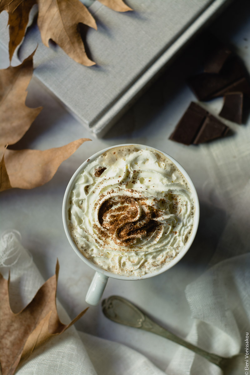 Homemade Pumpkin Spice Latte www.thefoodiecorner.gr 06 Photo description: A closer look at one of the cups of psl with whipped cream and a sprinkling of spices and shaved chocolate. Dry leaves to the left and the handle of an old silver spoon towards the bottom.