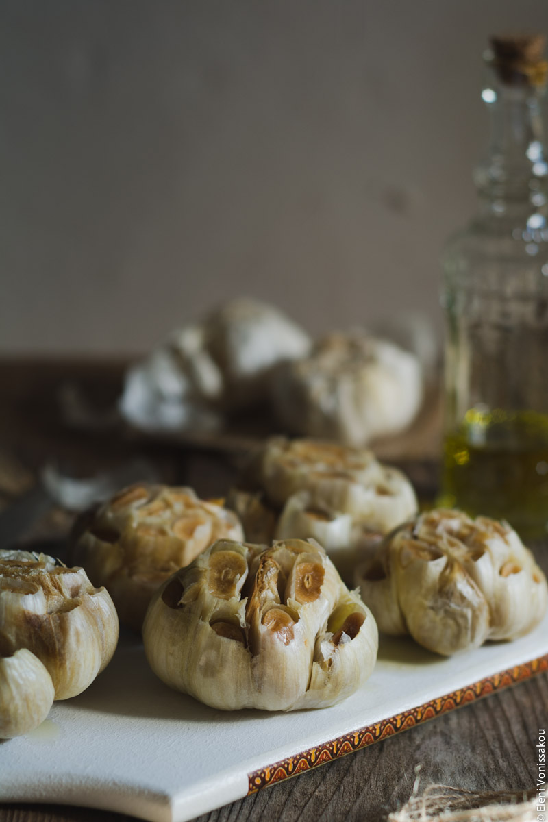 Ψητό Σκόρδο στο Slow Cooker www.thefoodiecorner.gr 03 Photo description: Side view of whole roasted garlic bulbs on a white chopping board with a pretty partern on the side. In the background a glass bottle of olive oil and some uncooked garlic.