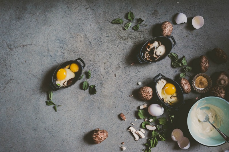 Food Styling and Photography Retreat – Crete, Greece 2017 www.thefoodiecorner.gr Photo description: Pictured to the right of the photo are some mini baking dishes with raw eggs in them, surrounding the dishes are mushrooms, eggs and greens. In the corner a bowl with a white sauce. All on a grey slate-like background.