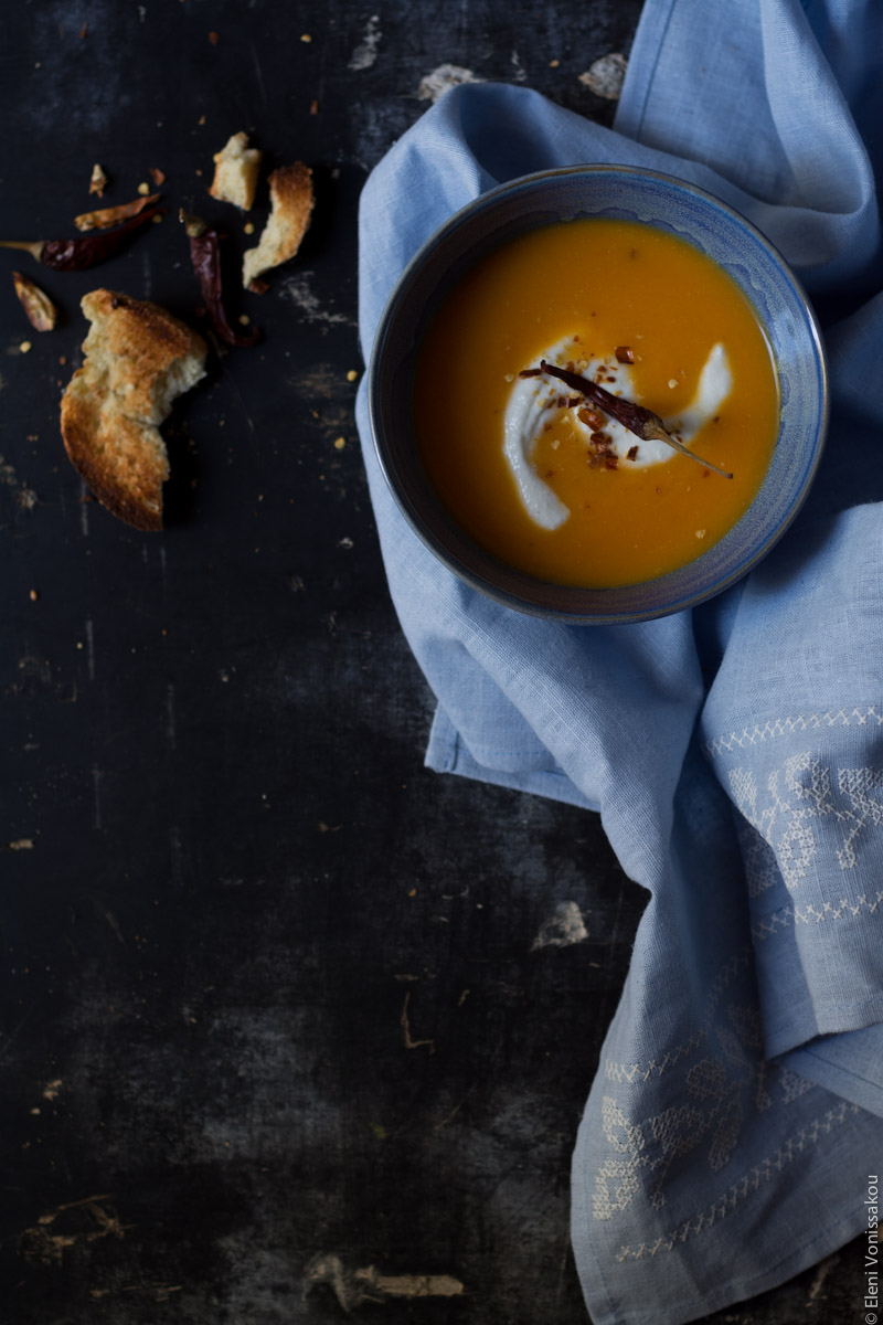 Butternut Squash Soup with Chilli and Whipped Goat’s Yoghurt Feta www.thefoodiecorner.gr Photo description: Overhead view of one blue bowl of butternut squash soup, in the top right corner of the frame, sitting on a light blue linen tea towel on a dark surface. The linen extends towards the bottom of the photo. On the soup is a swirl of whipped feta and a chilli pepper. To the left of the bowl is a broken piece of toasted bread.