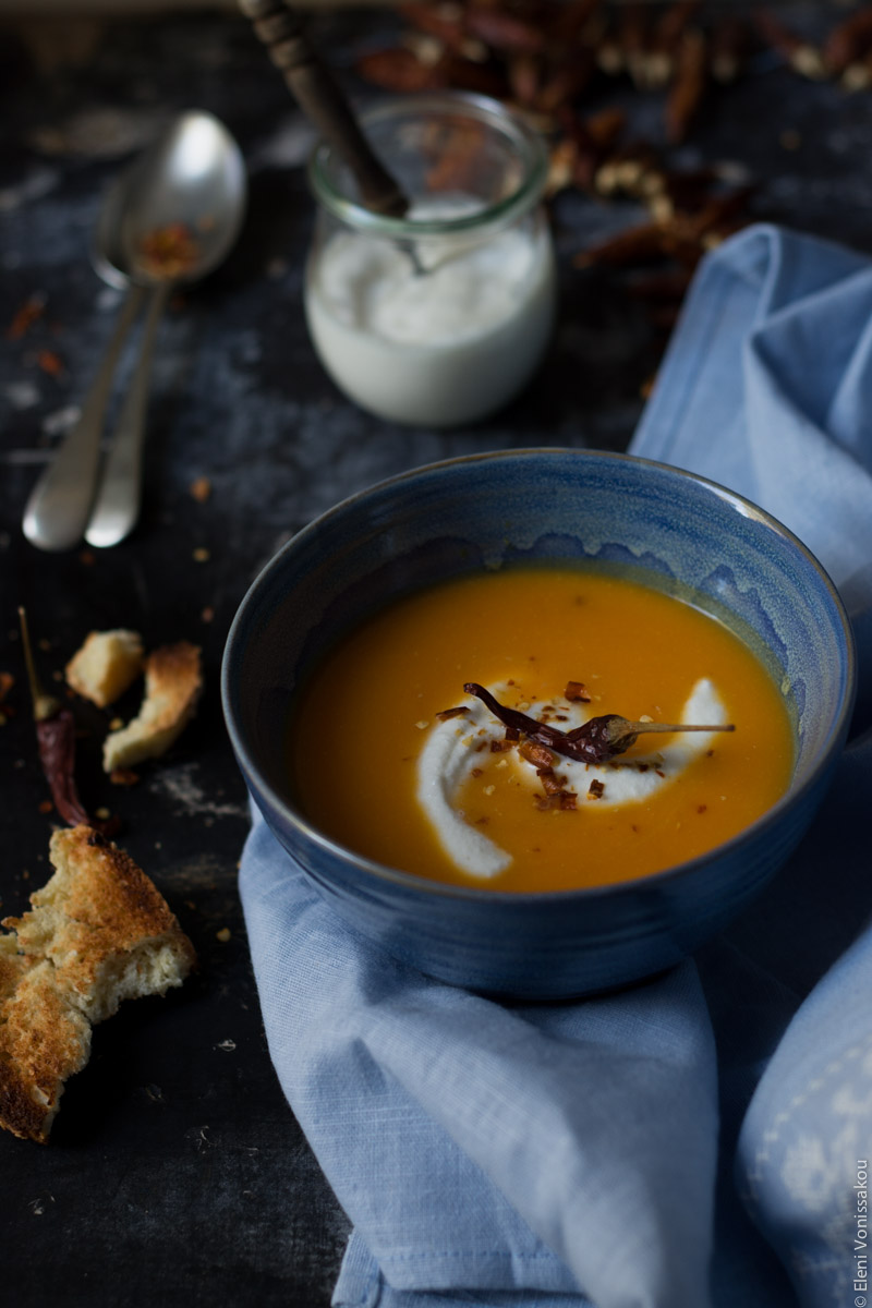 Butternut Squash Soup with Chilli and Whipped Goat’s Yoghurt Feta www.thefoodiecorner.gr Photo description: A 3/4 side view of one blue bowl of butternut squash soup, sitting on a light blue linen tea towel. A swirl of whipped feta and a chilli pepper sit on top of the soup. Behind the bowl is a small jar of whipped feta with a wooden handle spoon sitting in it. To the left of the bowl are some pieces of broken toasted bread. Right at the top a slightly blurred view of spoons and some whole dried chilli peppers.