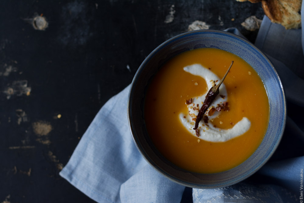 Butternut Squash Soup with Chilli and Whipped Goat’s Yoghurt Feta www.thefoodiecorner.gr Photo description: Overhead view of one blue bowl of butternut squash soup sitting on a light blue linen tea towel on a dark surface. On the soup is a swirl of whipped feta and a chilli pepper.