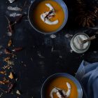 Butternut Squash Soup with Chilli and Whipped Goat’s Yoghurt Feta www.thefoodiecorner.gr Photo description: Overhead view of two bowls of butternut squash soup, one on each end of a dark surface. Around them along the edges are spoons, chilli flakes, whole dried chilli peppers and bits of toasted bread. In the bottom right corner is a light blue linen tea towel. On the right a small jar of whipped feta with a wooden handle spoon. On top of the bright orange soup is a swirl of whipped feta and a chilli pepper.