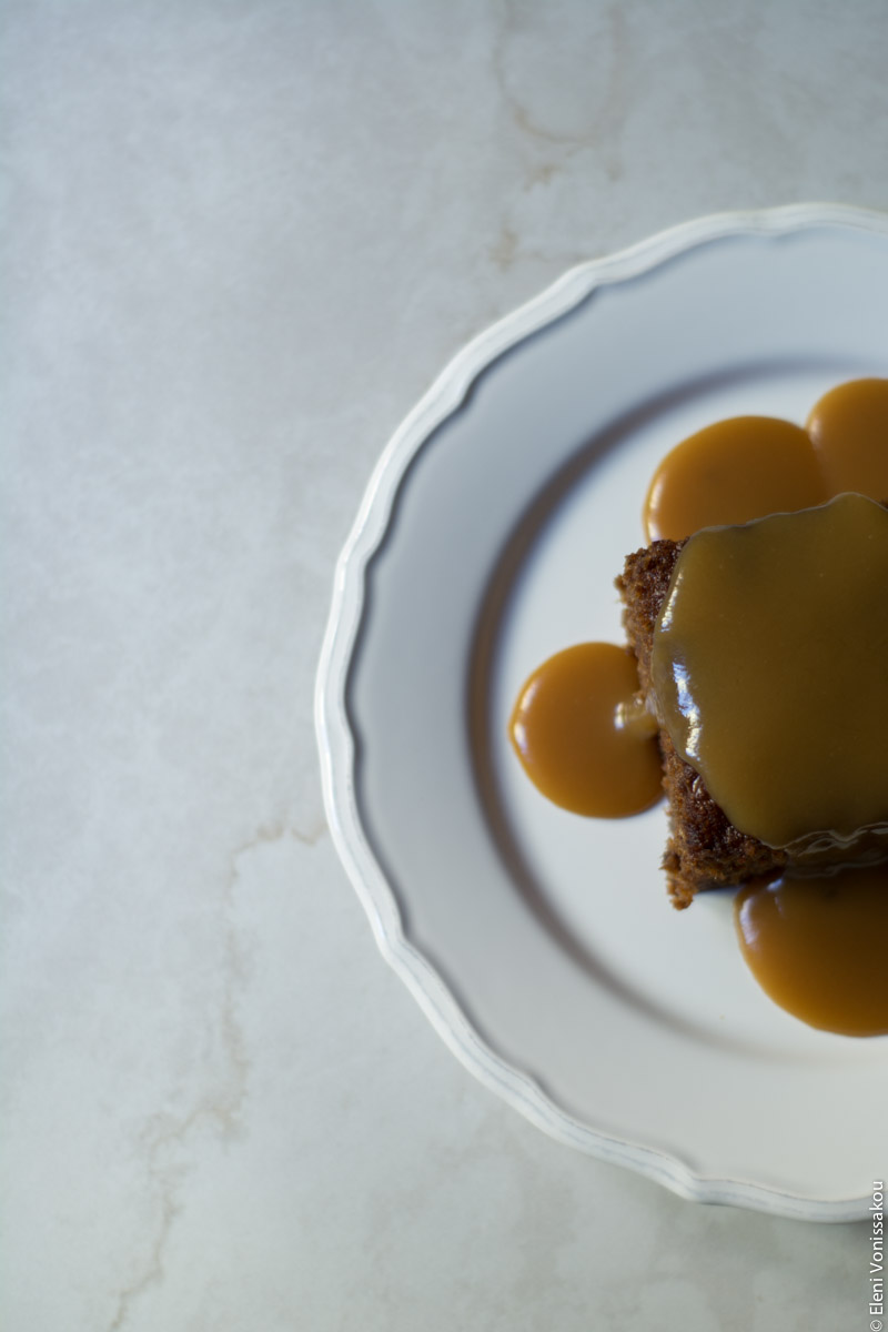 Chai Carob Sticky Toffee Pudding www.thefoodiecorner.gr Photo description: An overhead view of a piece of sticky toffee pudding with sauce all over it and down the sides, sitting on a white plate. Plate and pudding are to the side of the photo, half visible, sitting on a white marble-like surface.