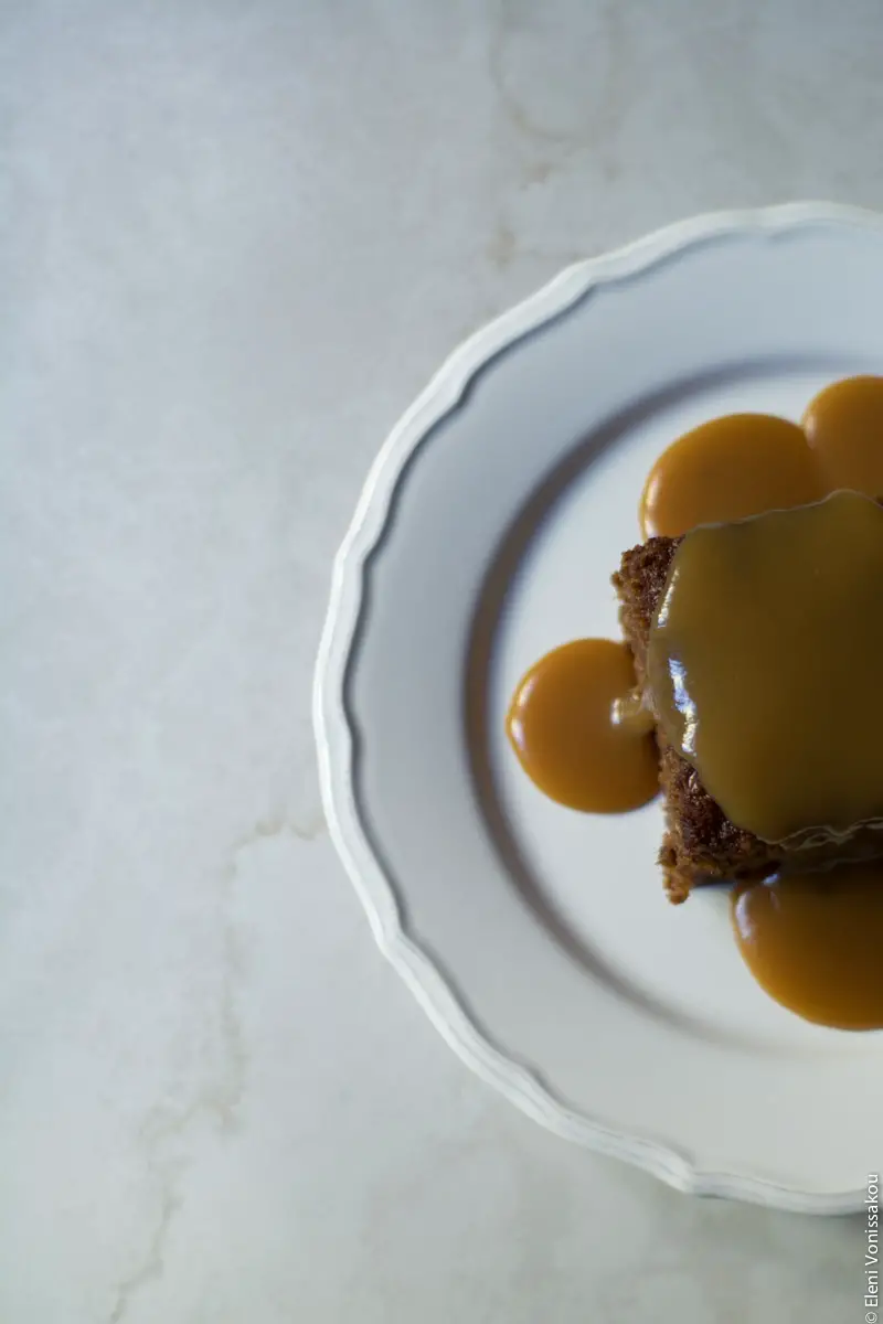 Sticky Toffee Pudding Κέικ με Χουρμάδες και Σάλτσα Καραμέλας Βουτύρου www.thefoodiecorner.gr Photo description: An overhead view of a piece of sticky toffee pudding with sauce all over it and down the sides, sitting on a white plate. Plate and pudding are to the side of the photo, half visible, sitting on a white marble-like surface.