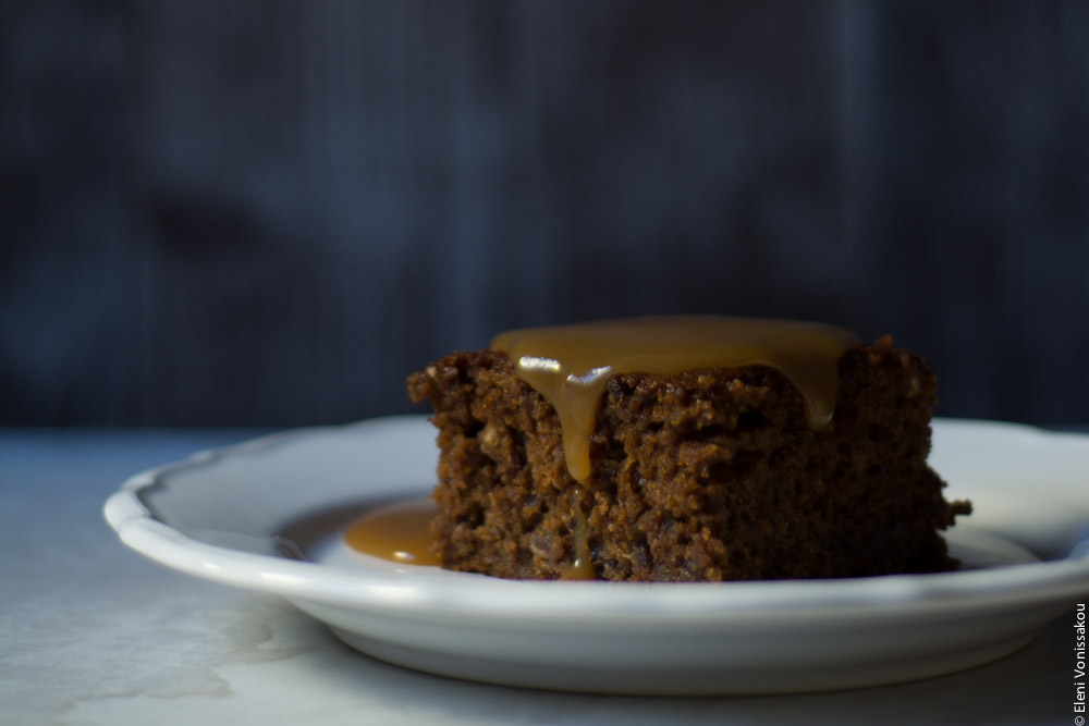 Chai Carob Sticky Toffee Pudding www.thefoodiecorner.gr Photo description: Side view of a plate with a piece of sticky toffee pudding on it, sauce running down the sides, in front of a dark background.