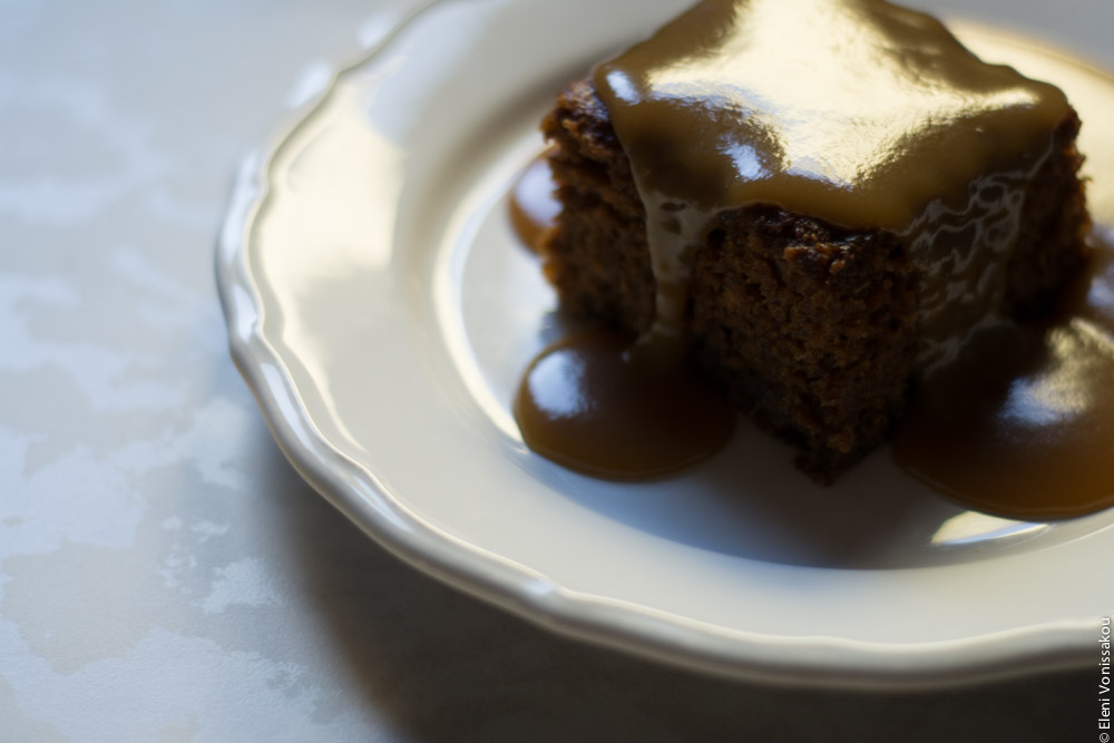 Chai Carob Sticky Toffee Pudding www.thefoodiecorner.gr Photo description: A piece of sticky toffee pudding with sauce all over it and down the sides, sitting on a white plate, light coming from behind darkening the front of the cake and making the sauce shine on top.