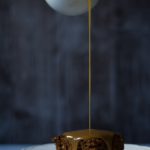 Chai Carob Sticky Toffee Pudding www.thefoodiecorner.gr Photo description: Side view of a plate with a piece of sticky toffee pudding on it, in front of a dark background, with a stream of sauce being poured over it, the jug just visible in the top of the photo. Sauce running down the sides of the pudding.