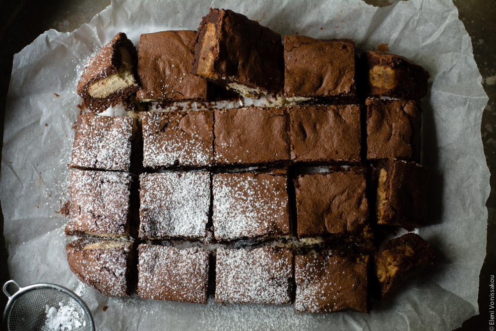 Brownies with Melomakarona and Kourabiedes - Aka Greek Christmas Brownies www.thefoodiecorner.gr The brownies on their baking paper, cut into squares and slightly separated. Half of them (on the diagonal) have been sprinkled with icing sugar. At the bottom left, barely visible, is the small sieve used to sprinkle the sugar.