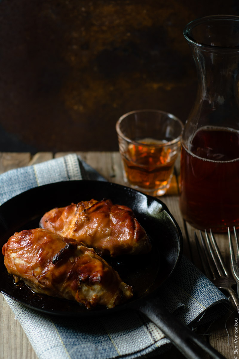 Prosciutto Wrapped Chicken Fillets with Greek Yoghurt, Mayo and Gruyere www.thefoodiecorner.gr Photo description: Side view of skillet with two prosciutto wrapped chicken breasts, a small glass and a jug of rose visible by its right side. Background is brown rust coloured.