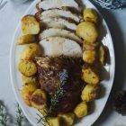 Slow Cooker Prosciutto Wrapped Turkey Breast with White Wine Gravy, and Rosemary Roast Potatoes www.thefoodiecorner.gr Photo description: Overhead view of turkey breast wrapped in prosciutto slices, laying on an oval serving platter with roast potatoes all around it. Half of it is sliced, the slices on their sides and overlapping. On top of the bottom half of meat lies a twig of flowering rosemary. Around the platter are Christmas decorations in silver and white colours. Surface is off white marble-like.