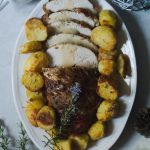 Slow Cooker Prosciutto Wrapped Turkey Breast with White Wine Gravy, and Rosemary Roast Potatoes www.thefoodiecorner.gr Photo description: Overhead view of turkey breast wrapped in prosciutto slices, laying on an oval serving platter with roast potatoes all around it. Half of it is sliced, the slices on their sides and overlapping. On top of the bottom half of meat lies a twig of flowering rosemary. Around the platter are Christmas decorations in silver and white colours. Surface is off white marble-like.