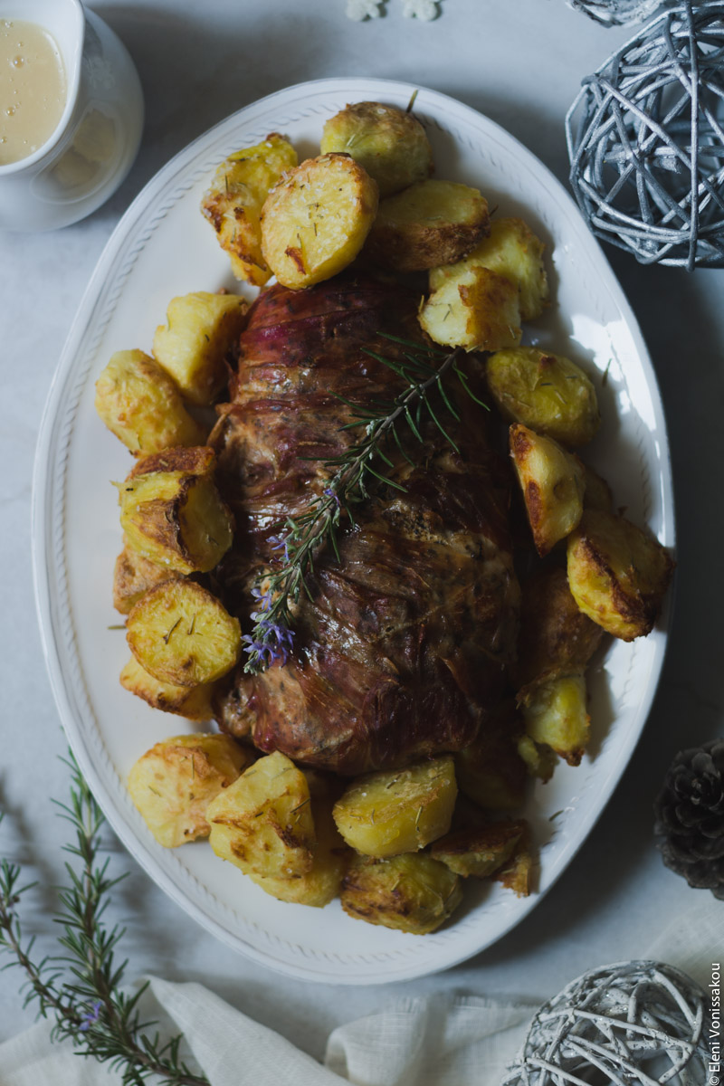 Slow Cooker Prosciutto Wrapped Turkey Breast with White Wine Gravy, and Rosemary Roast Potatoes www.thefoodiecorner.gr Photo description: Overhead view of turkey breast wrapped in prosciutto slices, laying on an oval serving platter with roast potatoes all around it. On top of the meat lies a twig of flowering rosemary. Around the platter are Christmas decorations in silver and white colours. Surface is off white marble-like.