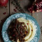 Slow Cooker Pomegranate and Tomato Beef with Sultanas www.thefoodiecorner.gr Photo description: Overhead view of a plate of beef over mashed potatoes with some pomegranate arils sprinkled on top. The antique plate has a pretty teal coloured design around the edge and is sitting on a wooden background. On the top side of the photo are some pomegranates, one of them broken open to reveal the arils inside. A couple of sprigs of thyme decorate the meat.