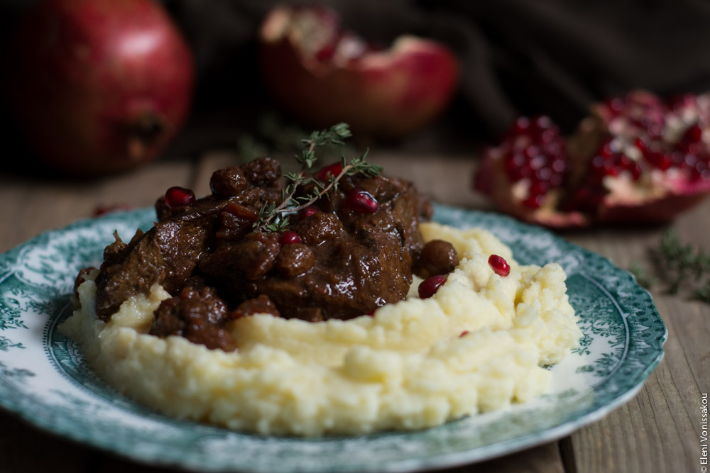 Slow Cooker Pomegranate and Tomato Beef with Sultanas www.thefoodiecorner.gr Photo description: Close up side view of a plate of beef over mashed potatoes with some pomegranate arils sprinkled on top. The antique plate has a pretty design around the edge and is sitting on a wooden background. Behind the plate are some pomegranates, one of them broken open to reveal the arils inside. A couple of sprigs of thyme decorate the meat.