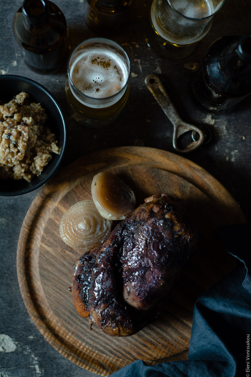 Slow Cooker Smoked Pork Knuckle (Ham Hock) with Beer and Jam www.thefoodiecorner.gr Photo description: Top view of a cooked ham hock on a round wooden cutting board. To its left towards the top of the photo is a black bowl with cooked bulgur wheat inside. Above the board are also some beer bottles and a couple of glasses with beer in. An old rusty bottle opener lies among them. In the bottom right corner of the photo lays a black linen napkin.