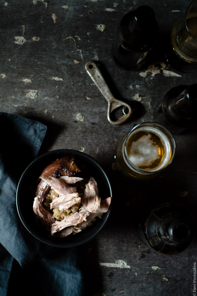 Slow Cooker Smoked Pork Knuckle (Ham Hock) with Beer and Jam www.thefoodiecorner.gr Photo description: A top view (portrait) of the bowl of bulgur wheat, with some torn pieces of ham hock laying on top, sitting in the bottom left corner. To its left a black linen napkin just visible, and to the right a glass of beer and the old rusty bottle opener. Towards the top of the photo on the left there is blank space showing the surface on which the items sit, which is dark and battered, with discoloured spots. To the right towards the top, some dark brown beer bottles.