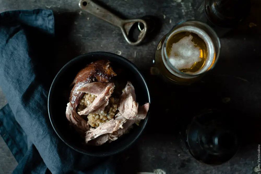 Slow Cooker Smoked Pork Knuckle (Ham Hock) with Beer and Jam www.thefoodiecorner.gr Photo description: A top view (landscape) of the bowl of bulgur wheat, with some torn pieces of ham hock laying on top. To the left a black linen napkin and to the right a glass of beer and the old rusty bottle opener.