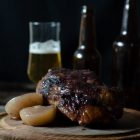 Slow Cooker Smoked Pork Knuckle (Ham Hock) with Beer and Jam www.thefoodiecorner.gr Photo description: Side view of a smoked ham hock sitting on a round wooden cutting board, with two cooked onion halves at the side. The hock is dark in colour and shiny. Behind it, a couple of dark brown beer bottles and one half full glass of beer. All set in front of a very dark and shadowy background.