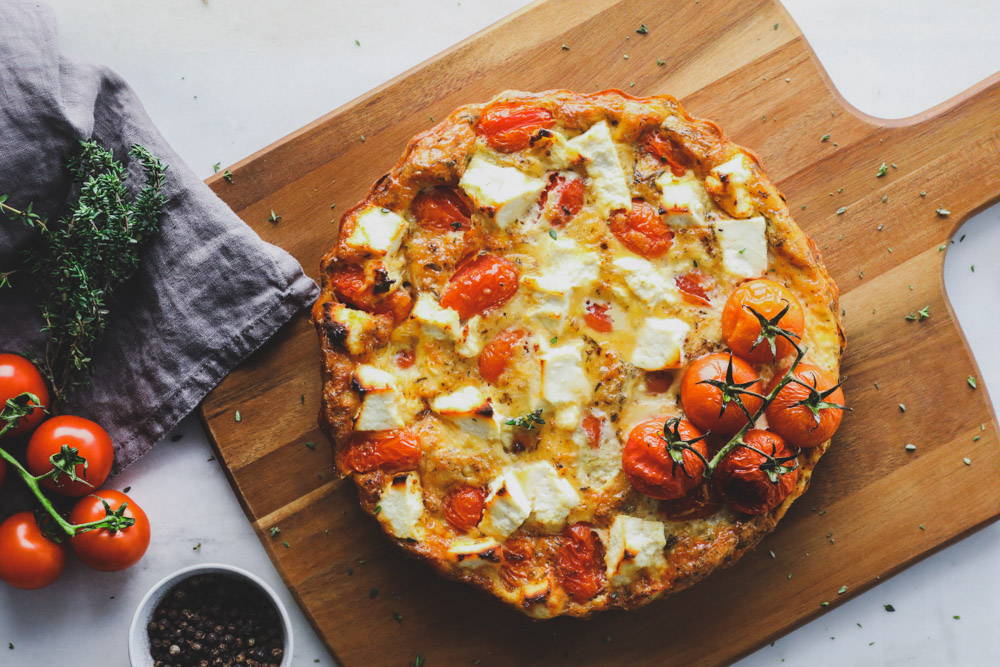 Easy Frittata with Spinach, Cherry Tomatoes and Anthotyro (Ricotta) www.thefoodiecorner.gr Photo description: A round frittata sitting on a wooden chopping board. The top is dotted with pieces of anthotyro and cherry tomatoes. At the side, a few cherry tomatoes on the vine, a linen tea towel and a bunch of thyme. A small bowl of pepper corns just visible at the bottom of the photo.