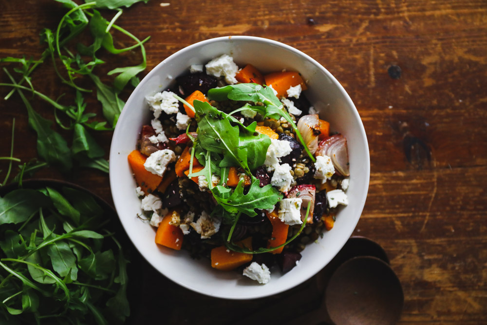 Warm lentil salad with roast beetroot, pumpkin and goat’s feta www.thefoodiecorner.gr Photo description: Top view of a bowl of lentil salad, pieces of beetroot, pumpkin, onion and feta scattered all over. Some rocket leaves garnishing the top. Bowl is sitting on a dark wooden surface, with more rocket leaves scattered to its left. Wooden serving spoons just visible to the right of the bowl.