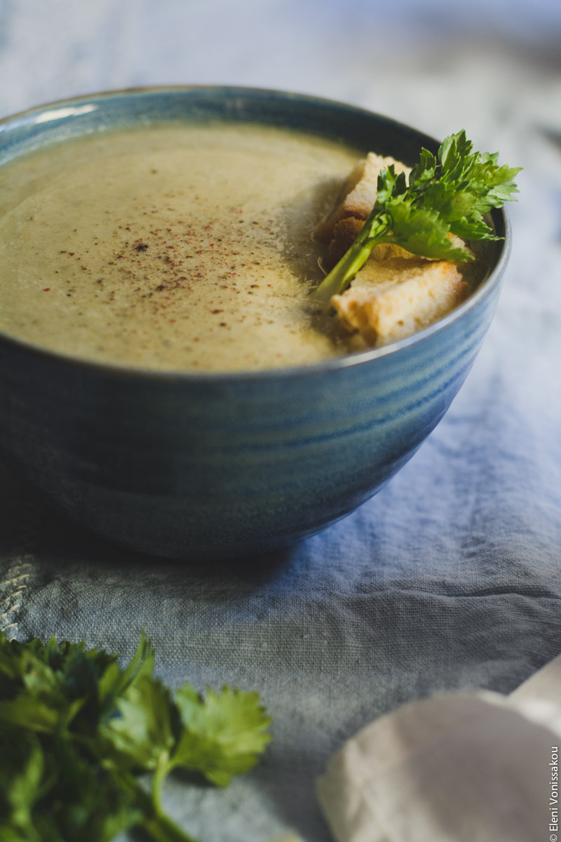 Slow Cooker Cream of Celery Soup with Mascarpone www.thefoodiecorner.gr Photo description: A close up three quarter view (portrait orientation) of one of the bowls of soup, sitting on a light blue linen cloth. In front and to the right, a corner of a white linen napkin is just visible and to the left some celery leaves. Some croutons and a small celery stalk garnish the soup on one side of its surface.