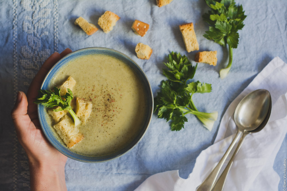 Slow Cooker Cream of Celery Soup with Mascarpone www.thefoodiecorner.gr Photo description: A single bowl of soup on a light blue linen cloth. To the left of the bowl, my hand holding the side. To the right, some scattered celery leaves and croutons. Further right a white napkin with two spoons sitting on it.