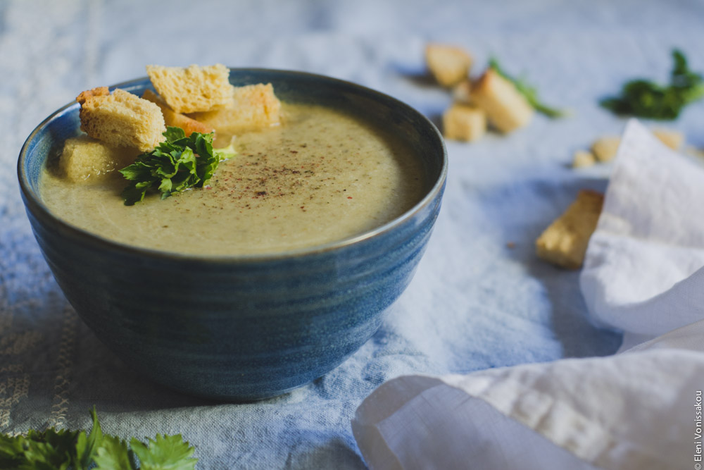 Slow Cooker Cream of Celery Soup with Mascarpone www.thefoodiecorner.gr Photo description: A close up three quarter view of one of the bowls of soup, sitting on a light blue linen cloth. To its right, a bunched up white linen napkin and some scattered croutons and celery leaves. Some croutons and a small celery stalk garnish the soup on one side of its surface.